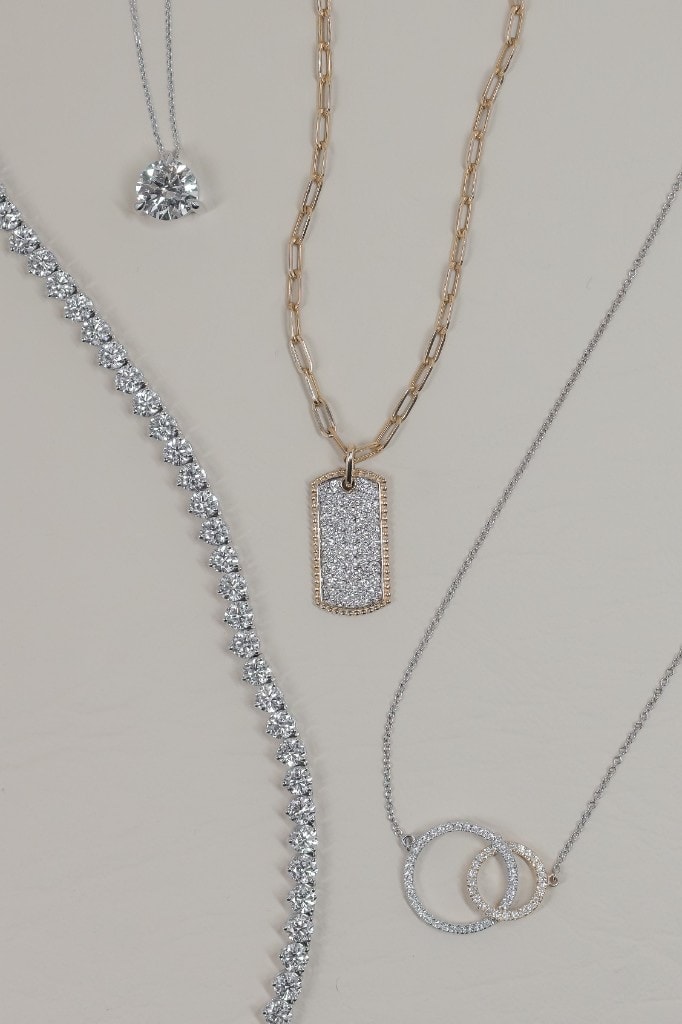 Layered diamond necklaces — a solitaire pendant, tennis necklace, pavé dog tag on a gold paperclip chain, and interlocking diamond circles