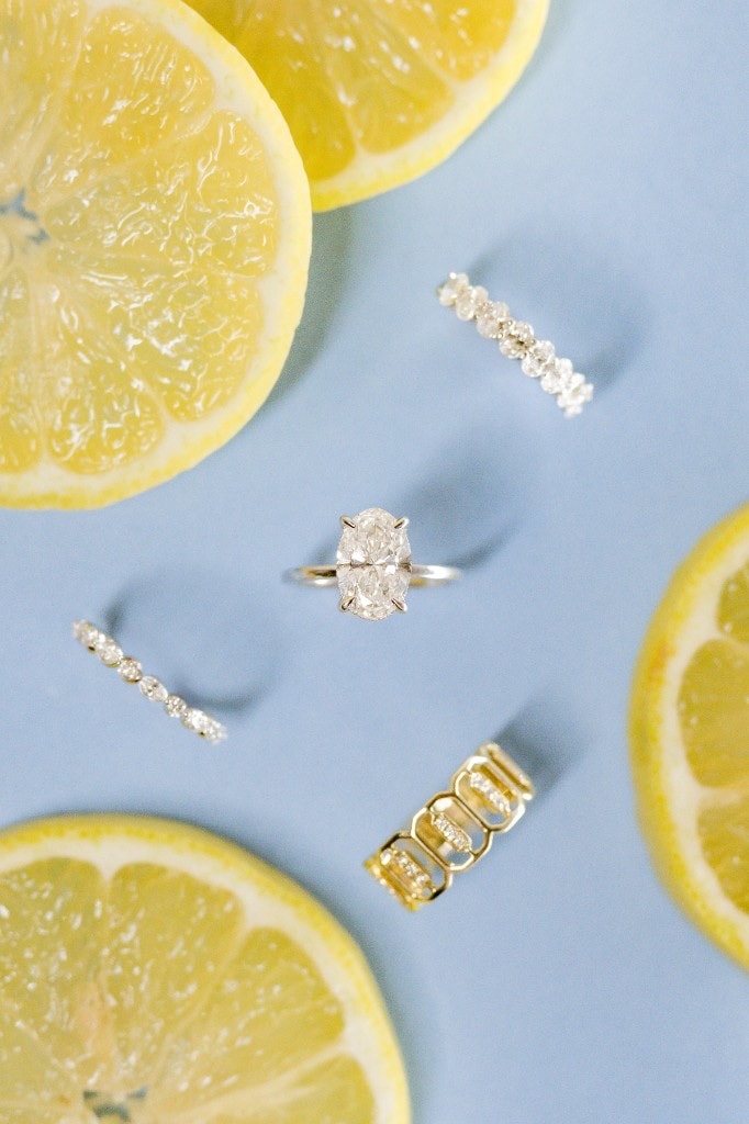 Oval solitaire engagement ring and diamond eternity bands styled with lemon slices on a powder blue background