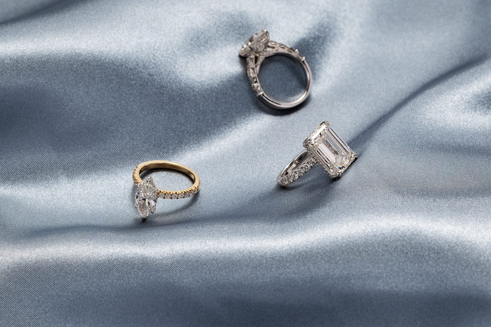 Three engagement rings on dusty blue silk: yellow gold marquise solitaire, marquise diamond halo, and emerald cut on a pavé band