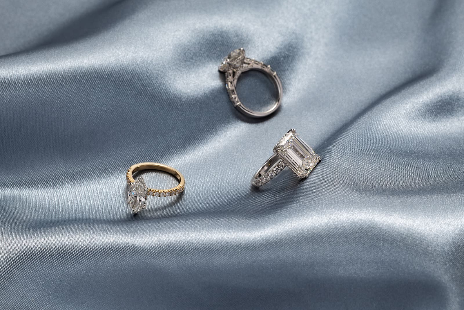Three engagement rings on blue silk — emerald cut, marquise, and round halo