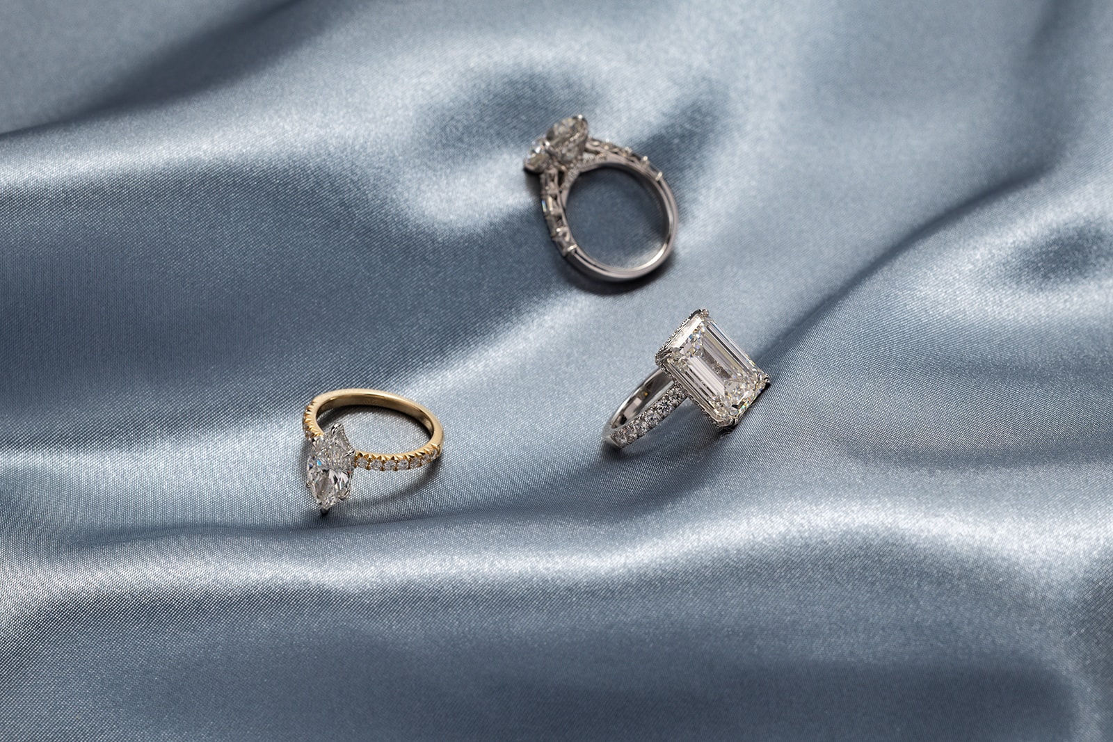 Three engagement rings on blue silk: emerald cut, marquise, and round halo