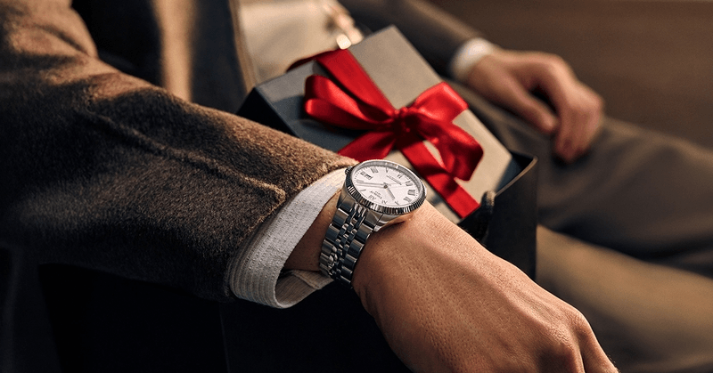 Tissot watch worn on the wrist beside a gift box tied with a red ribbon