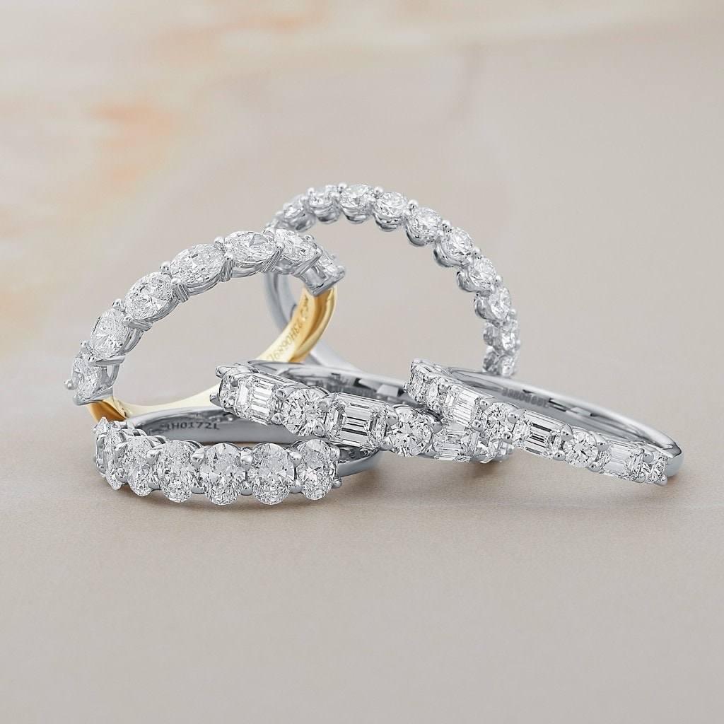 Diamond eternity bands with oval, round, marquise, and emerald-cut stones leaning in a loose stack on cream marble