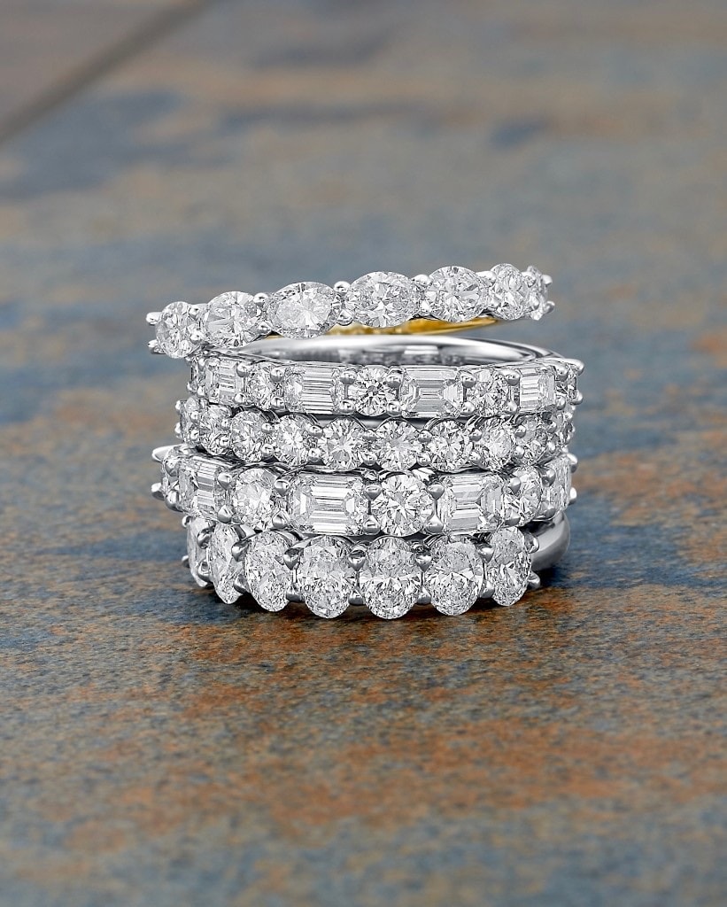 Five diamond wedding bands stacked together on slate, mixing oval, round, and emerald-cut diamonds in white gold