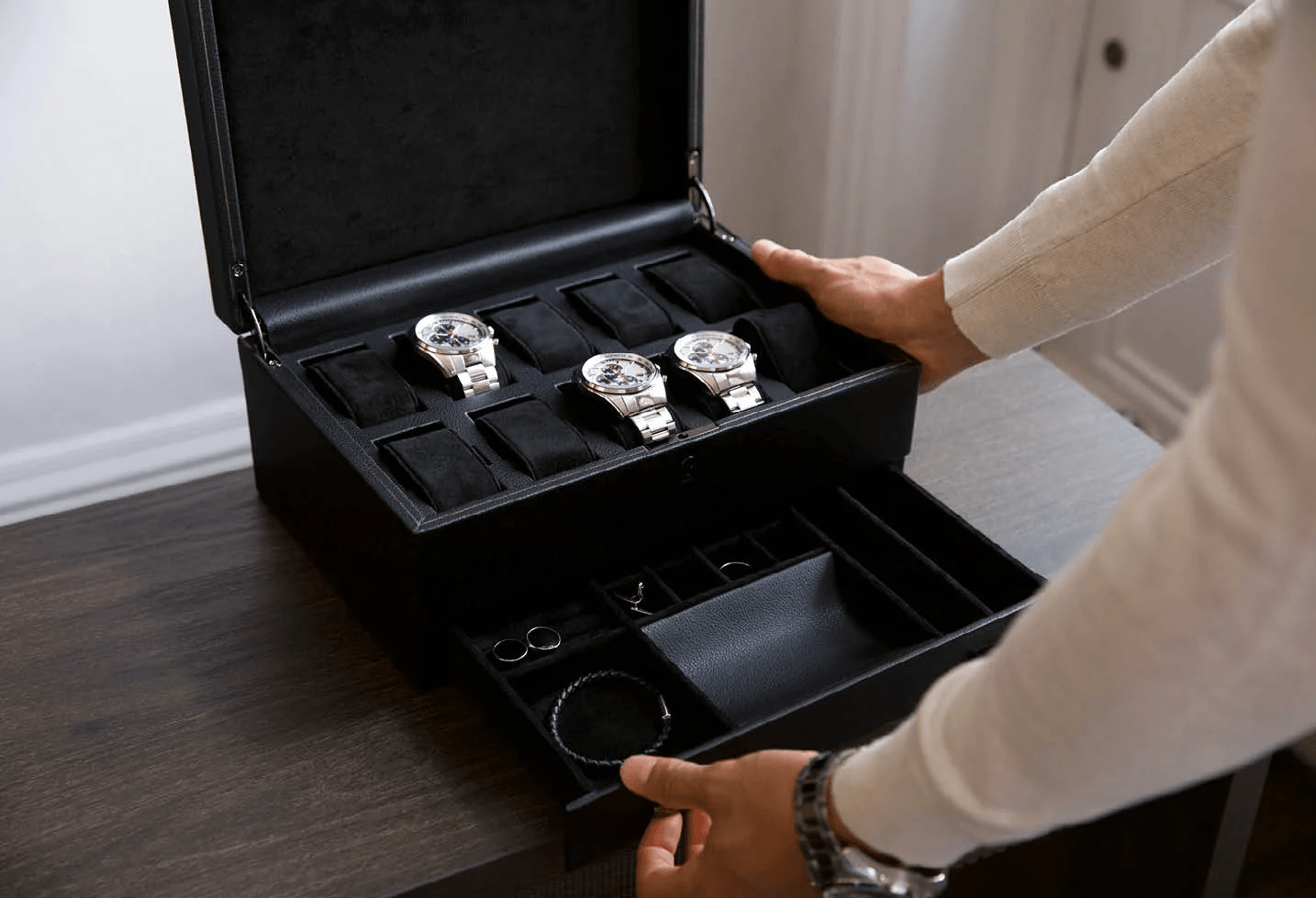 Open black watch box holding three watches with an accessory storage drawer